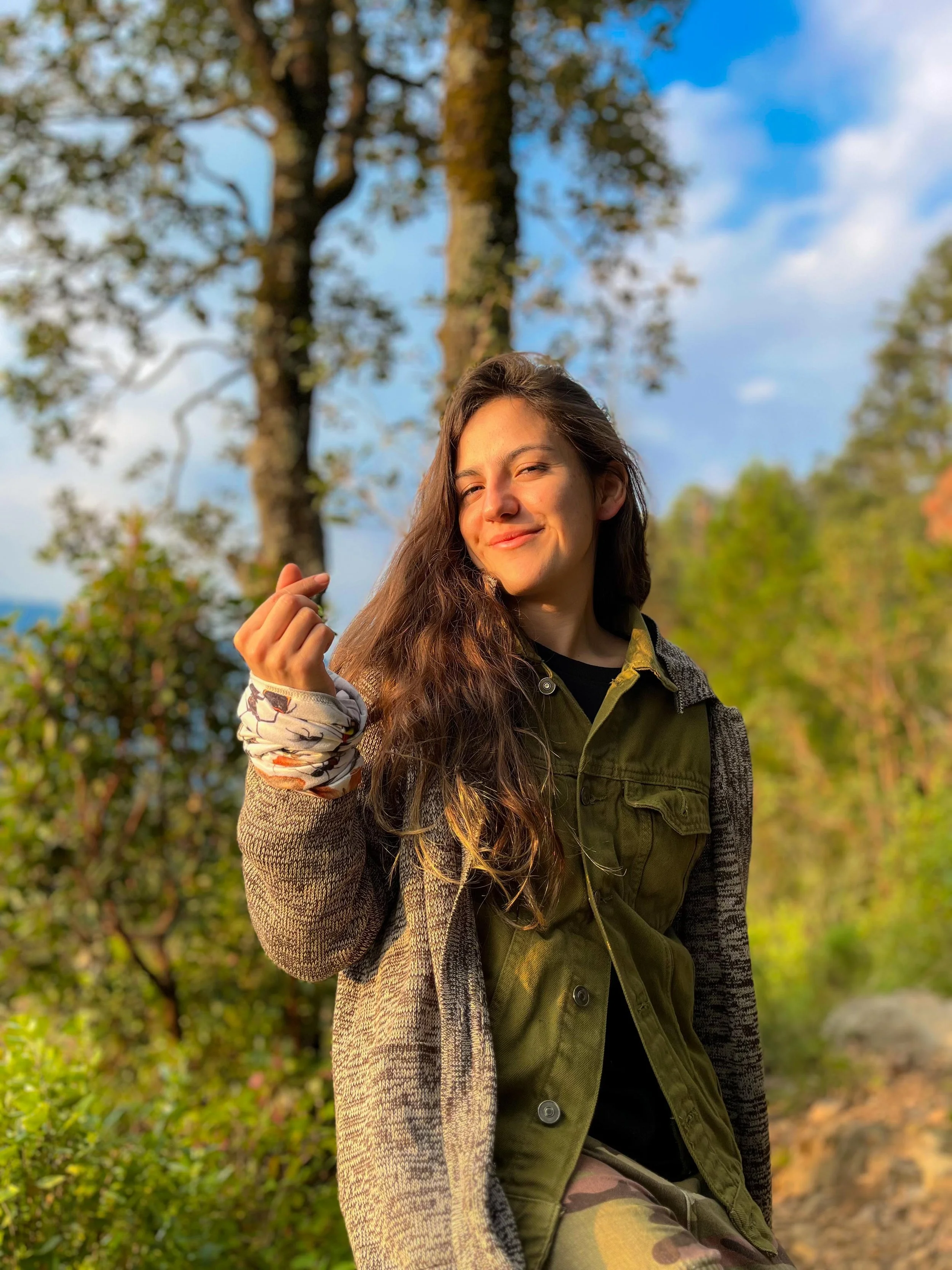 Biol. Dulce Díaz , Guia de excursionismo de AlMonte Hike.  Joven mujer con cabello rizado y sonrisa, sosteniendo una flor, en un entorno natural con árboles de fondo y cielo azul con nubes.