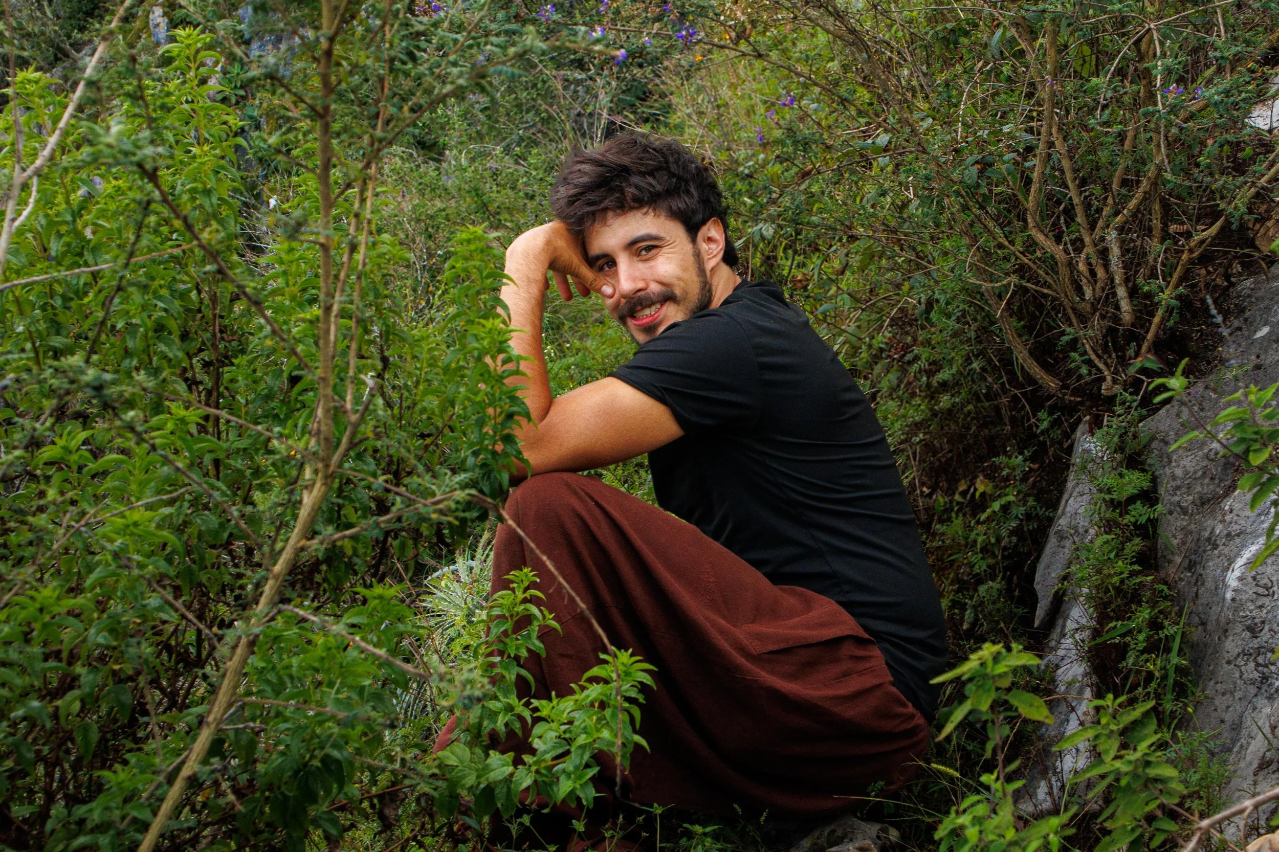 Un hombre sentado entre vegetación frondosa, con cabello oscuro y barba, sonriendo y descansando en un entorno natural.