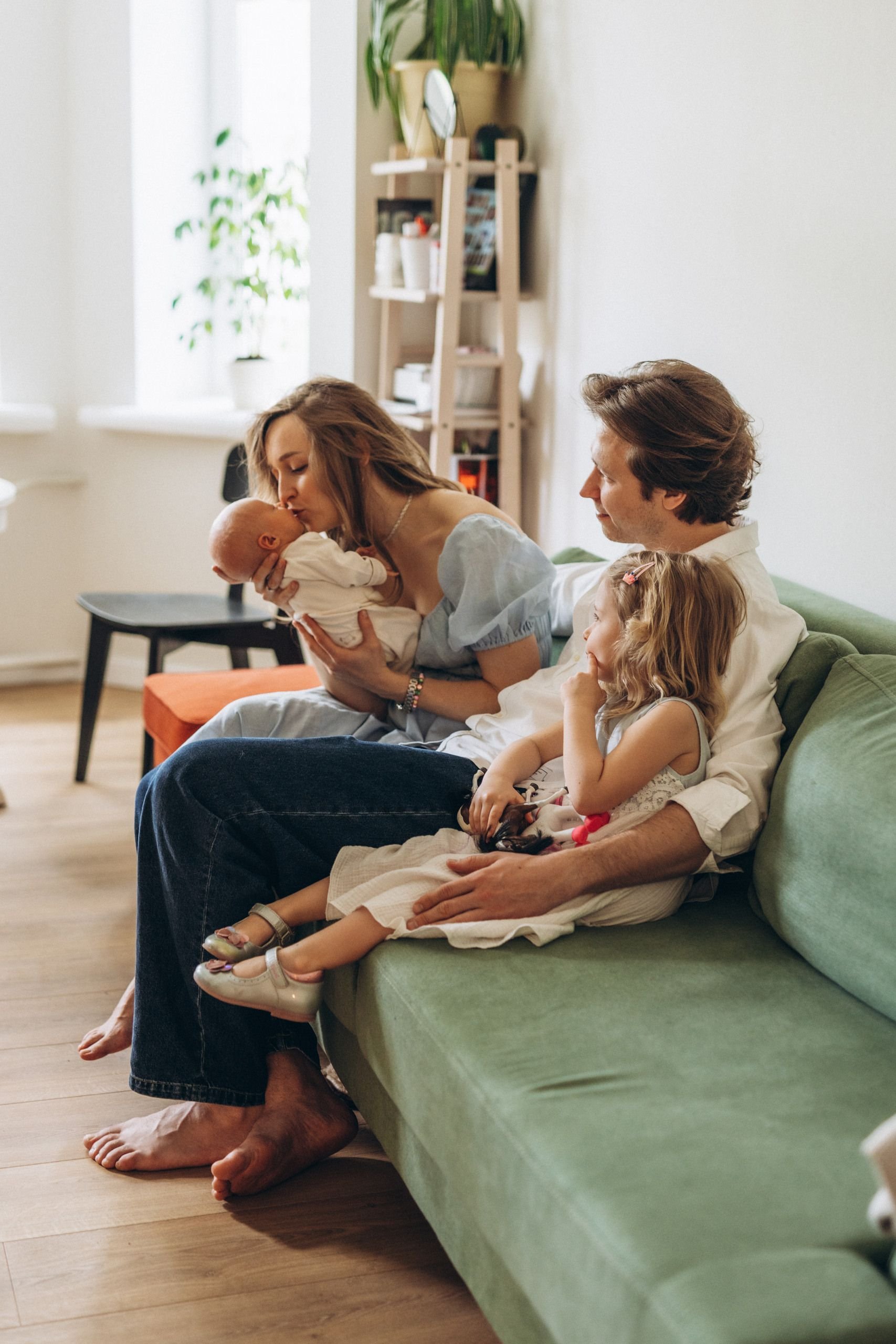 A family sitting on a green couch in a living room, with a woman kissing a baby, a man holding a young girl, and a child leaning on the woman. There is a ladder-style bookshelf, a window, and a small black table in the background.