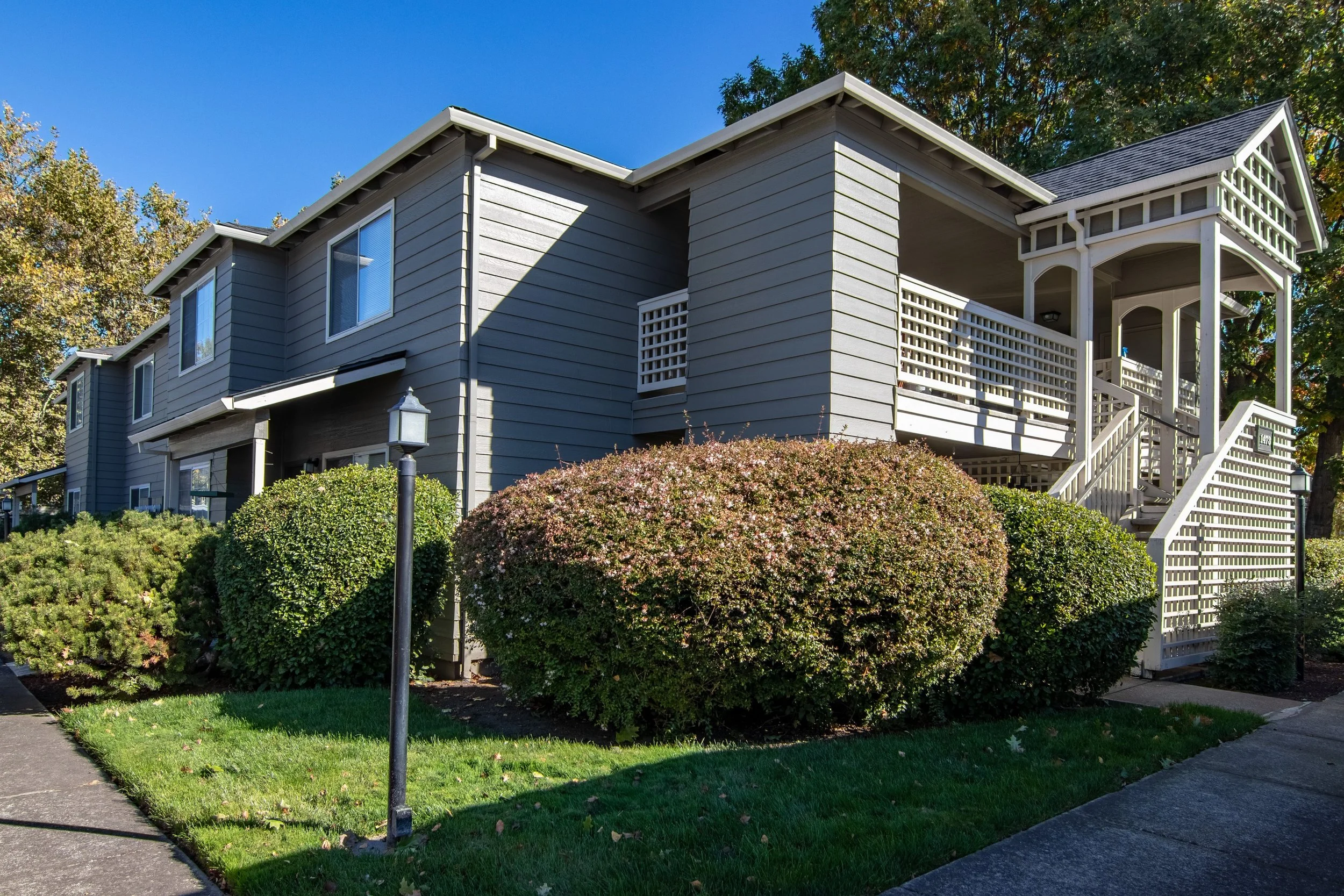 A gray multi-story residential building with a covered porch, white lattice details, and stairs, surrounded by green shrubs, trees, and a lamp post against a clear blue sky.