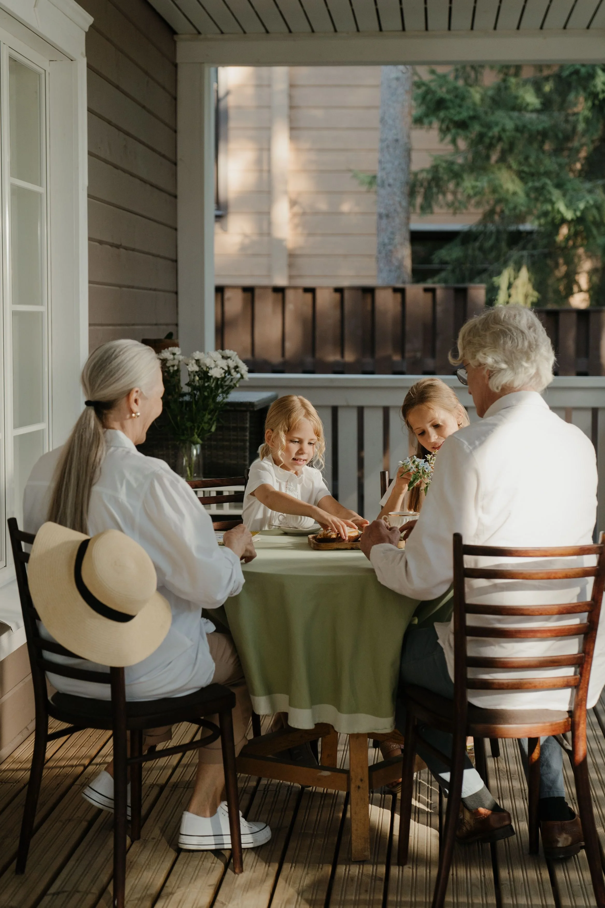 Three generations of a family, including children, grandparents, and a mother, sitting around a table on a porch, sharing a meal. The porch has wood paneling, a window, and a lush green tree in the background.