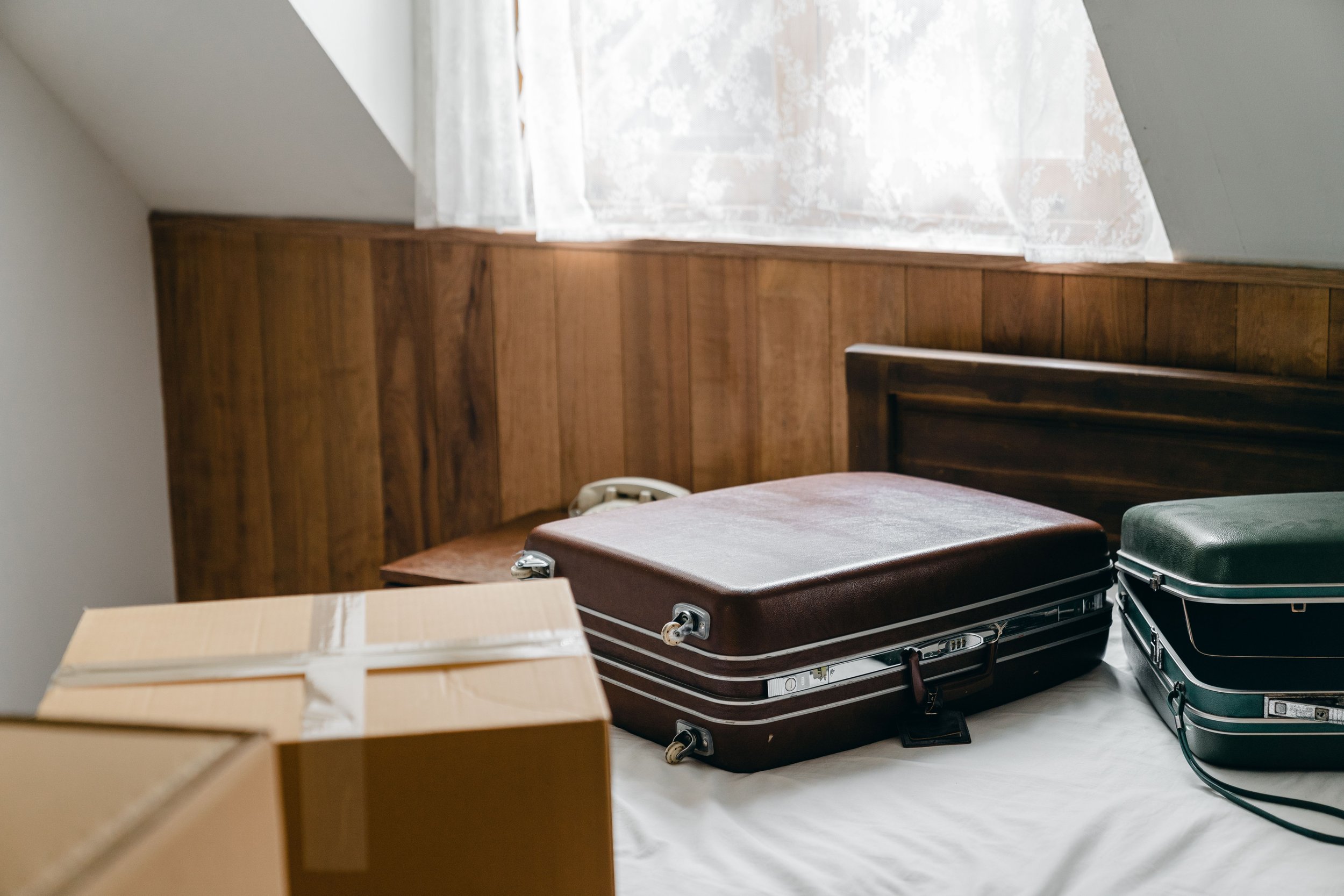 A corner of a room with a wooden wall panel, a window with lace curtains, a cardboard box, and three vintage suitcases on the floor.