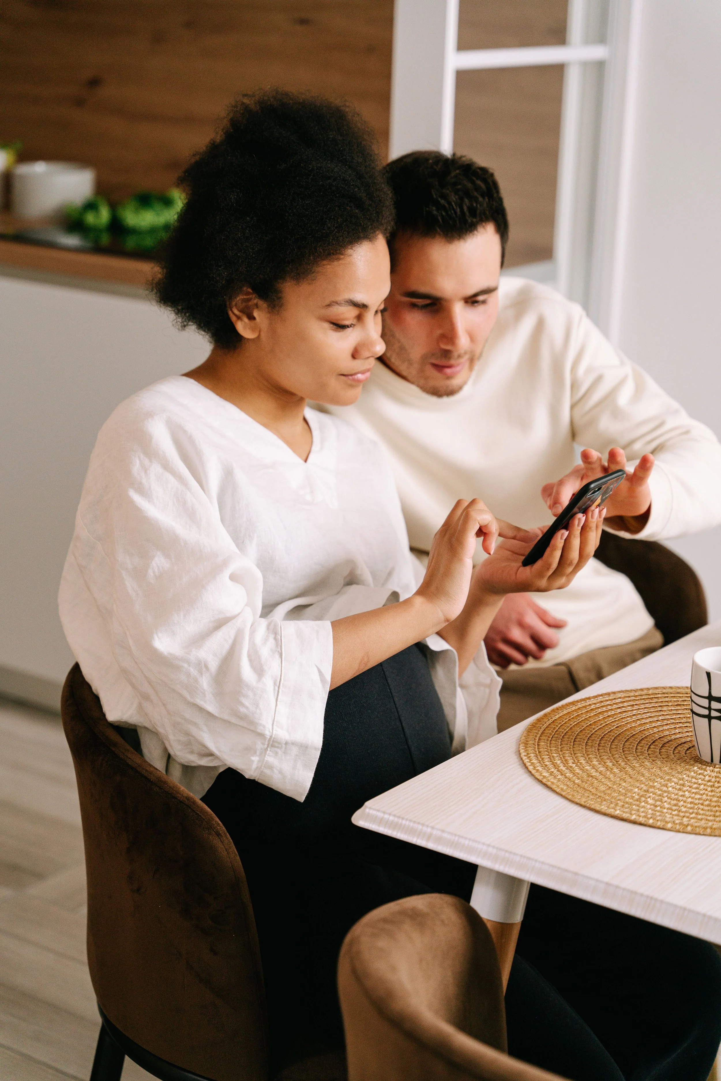 A woman and a man sitting at a table, looking at a smartphone together in a cozy kitchen setting.