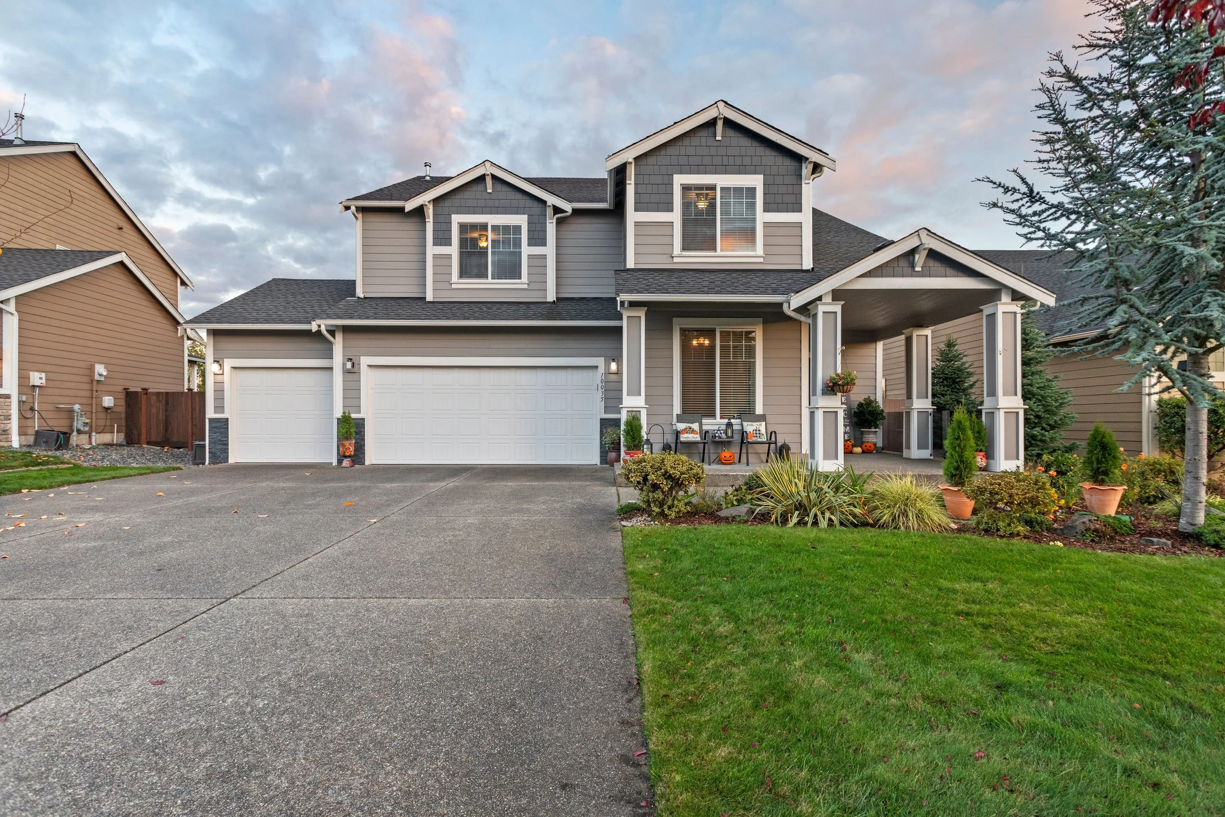 A modern two-story house featuring gray and beige siding, white trim, a nested garage with white doors, a porch with potted plants, and autumn-themed decorations outside. The front yard has a green lawn, bushes, and a large tree on the right, with a driveway leading up to the garage. The sky is partly cloudy at dusk.