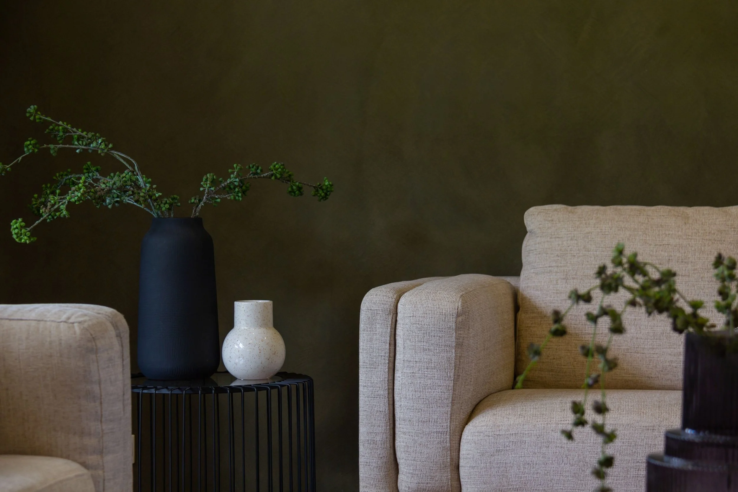 A beige armchair, a black vase with green plants, and two vases, one white and one with black stripes, on a black wire side table against a dark green wall.