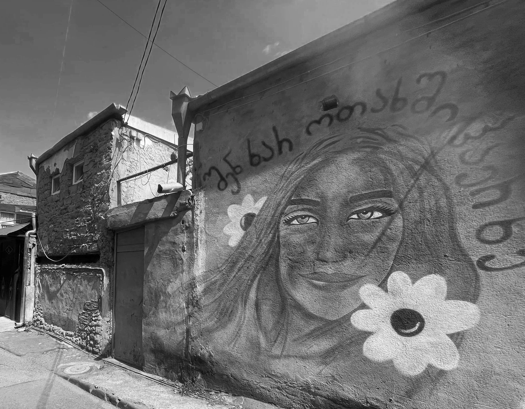 A large mural of a woman with long hair painted on the side of a building, with African-American features and expressive eyes. The building has textured, stone and stucco walls, with visible utility wires and pipes. There is graffiti or writing in a different language above the mural.