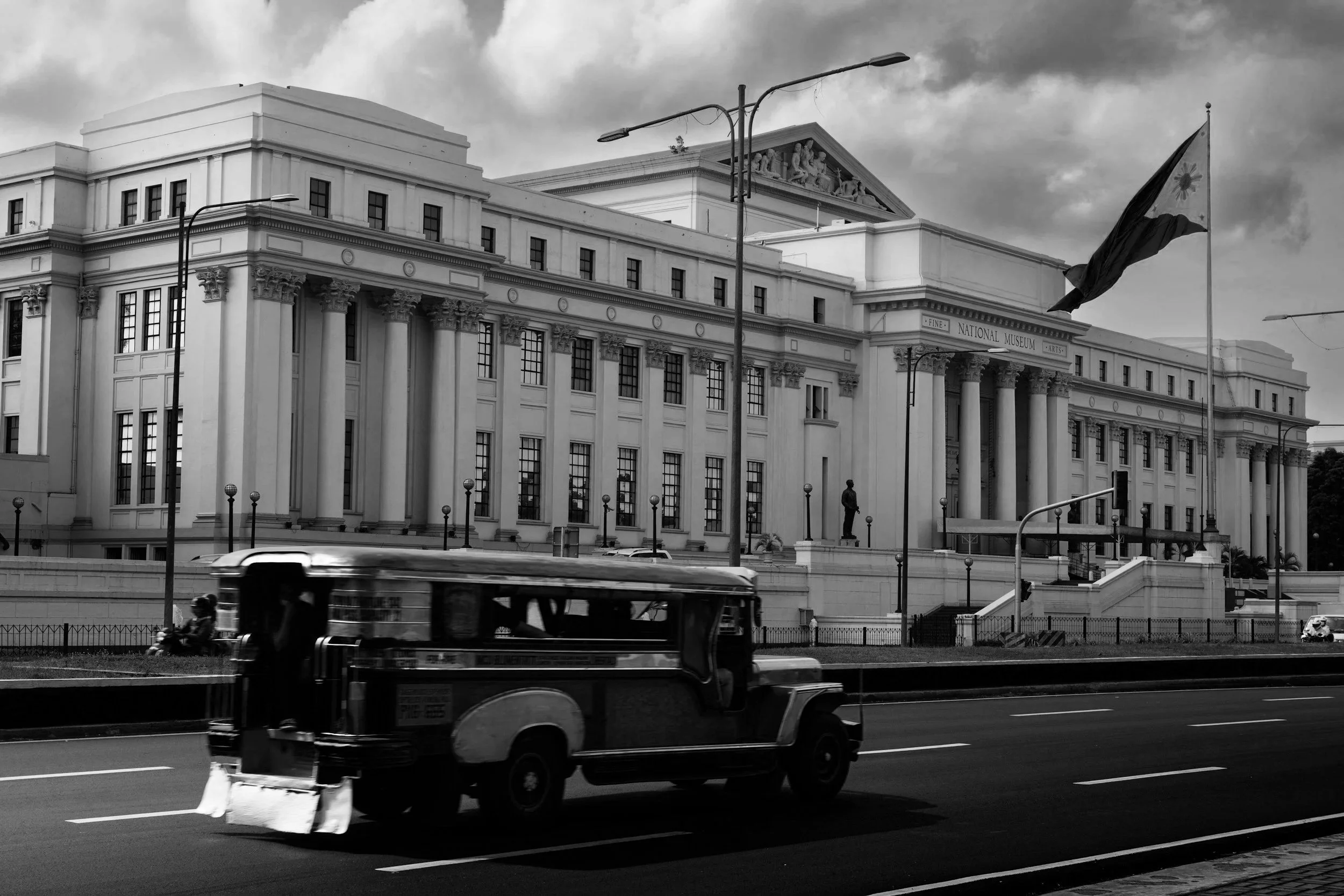 Black and white photo of a classical government building with columns and a pediment featuring sculptures. A street scene in front includes a moving bus, a motorcycle with a rider, and an overhead streetlight. A flag is visible on the right side of the image.
