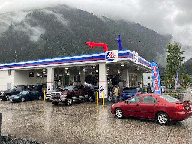 A gas station with a blue, red, and white canopy, and an inflatable dinosaur on top. Several cars are parked and being refueled. Mountains and cloudy sky in the background.