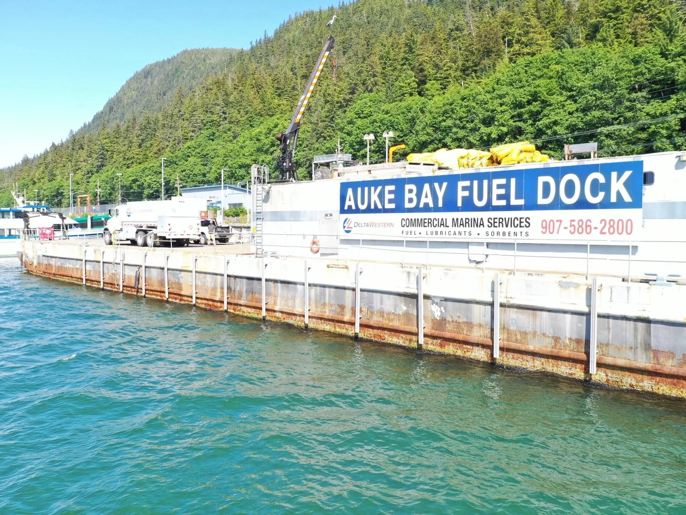 A dock with a sign that reads 'Auke Bay Fuel Dock'; there are trucks and equipment on the dock, with a green, forested hillside in the background.