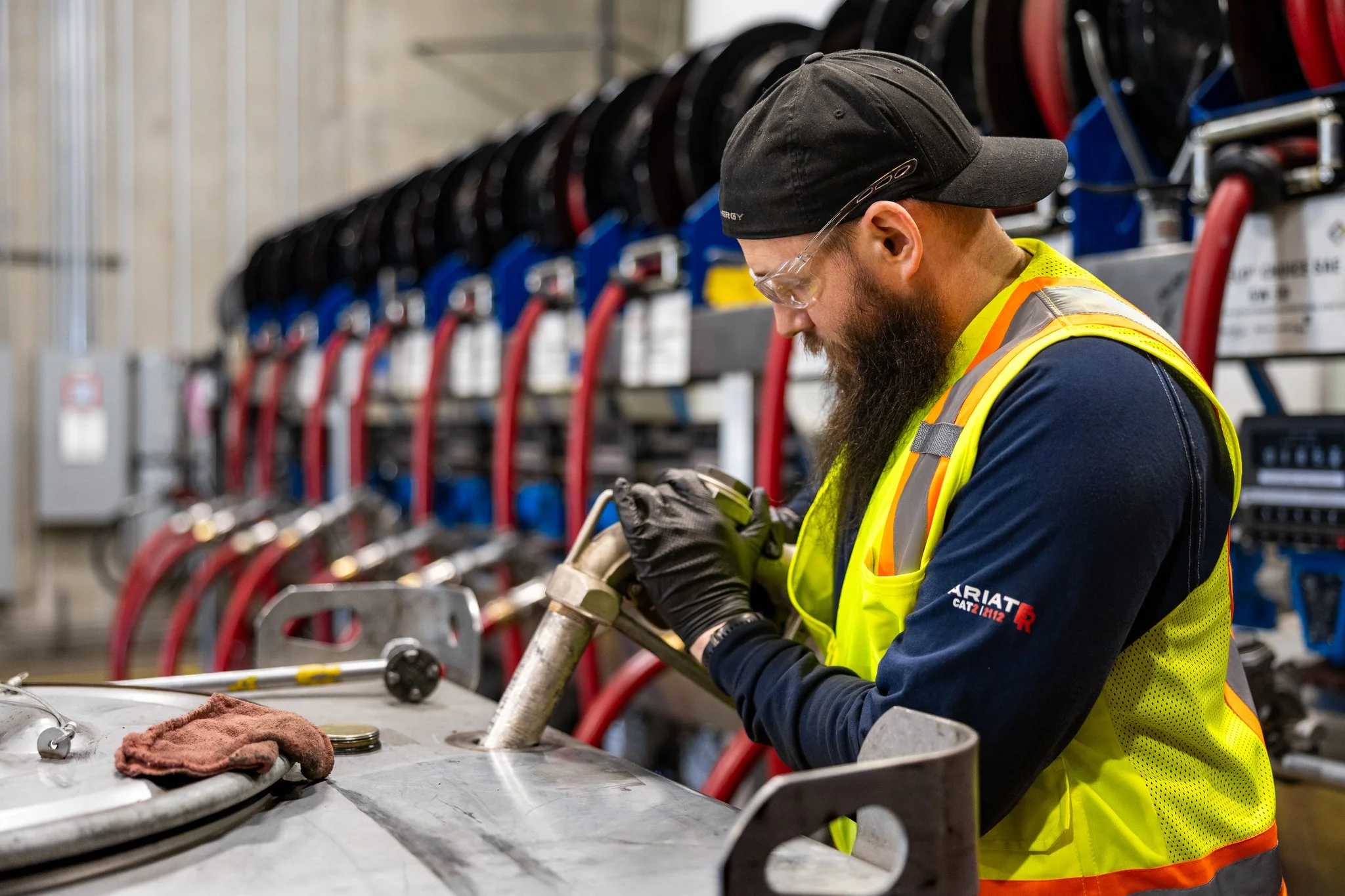 A worker wearing safety glasses, a black cap, and a yellow safety vest is working on a metal pipe in an industrial setting. There are tools and equipment on the workbench and a rack of helmets in the background.