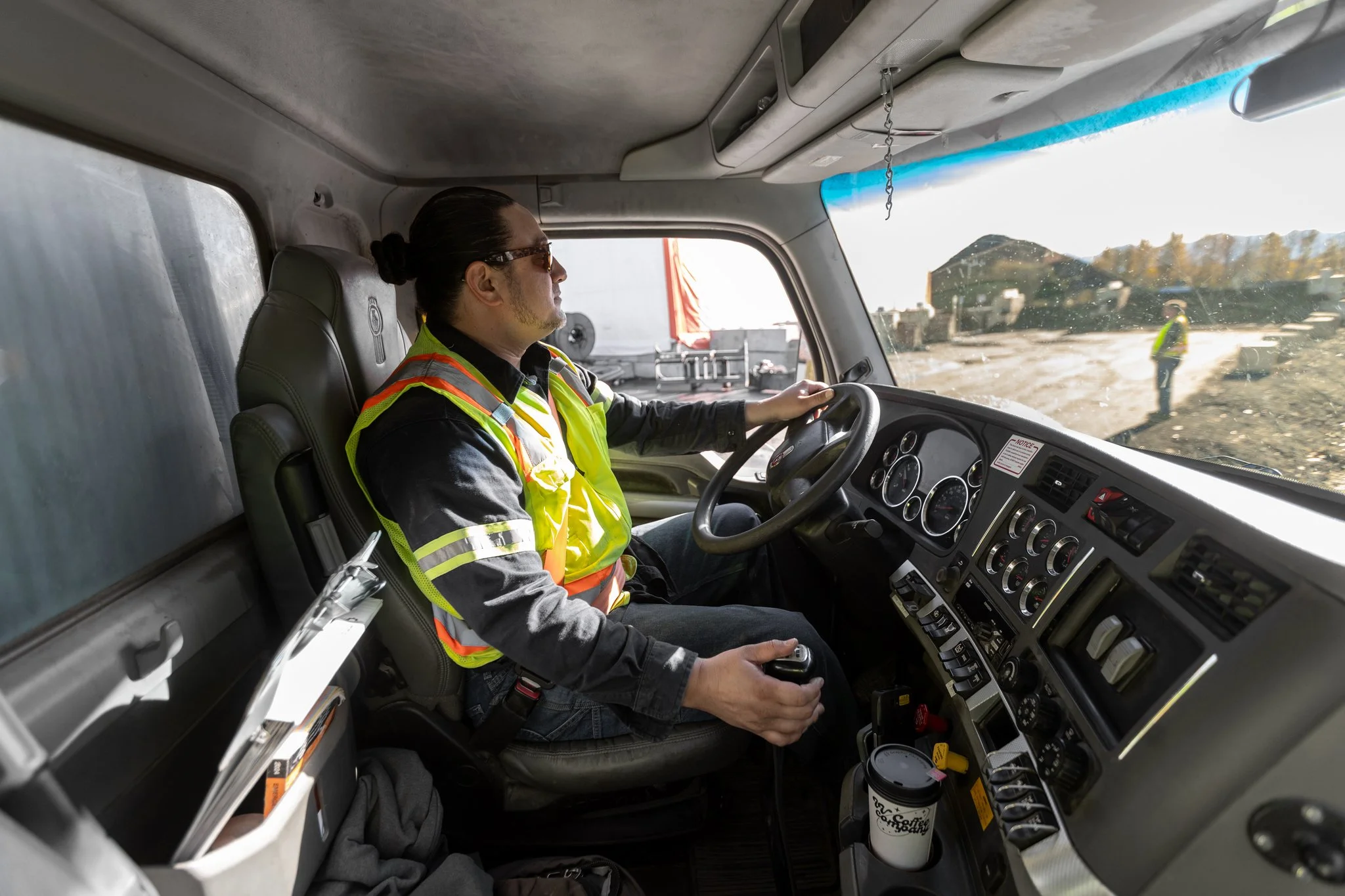 A man with dark hair tied in a bun, wearing sunglasses and a high-visibility safety vest, is sitting in the driver's seat of a large vehicle, likely a truck, with a dirt road and construction worker visible outside through the windshield.