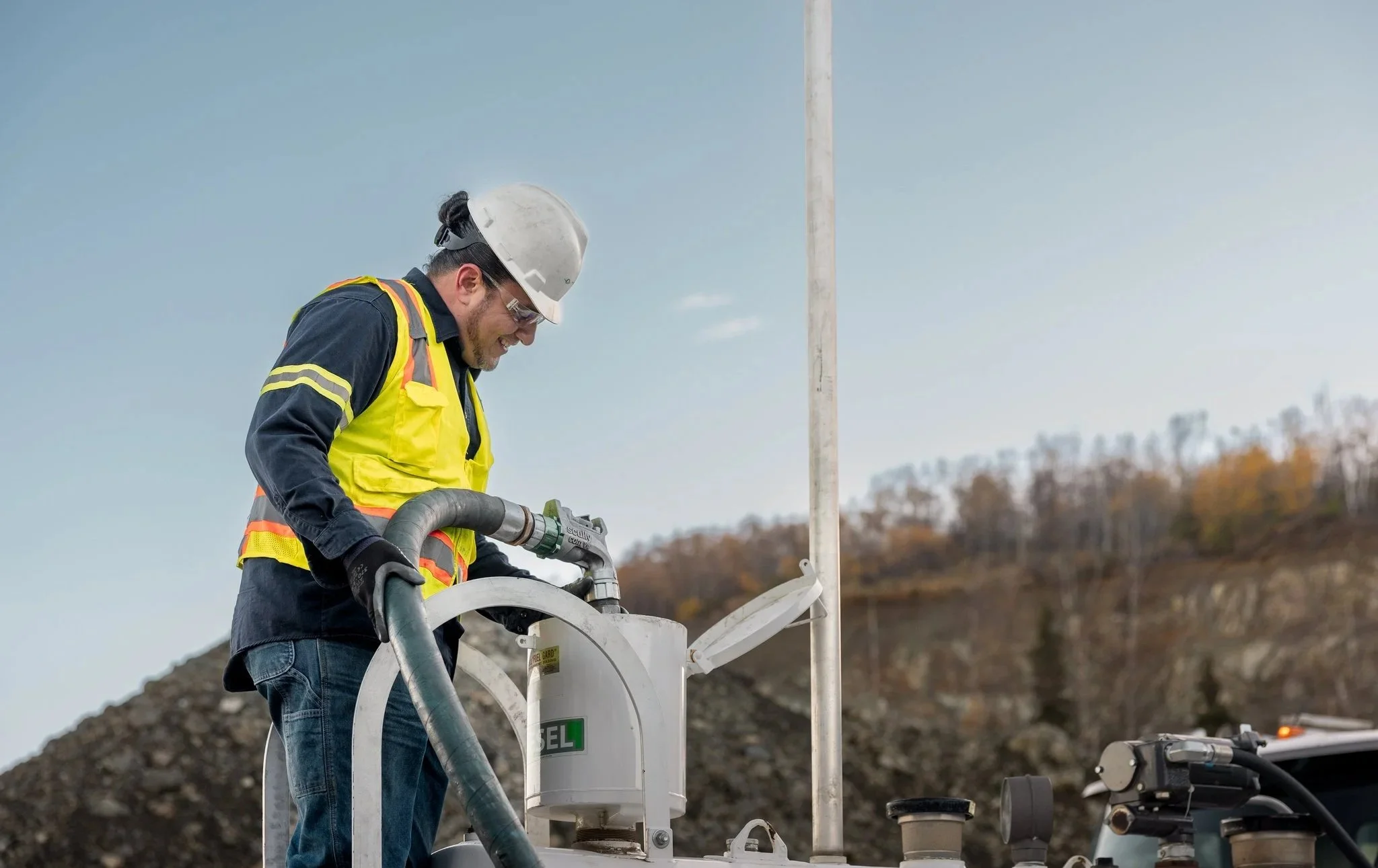 A male worker in safety gear operates equipment outdoors near a pole with hills in the background.
