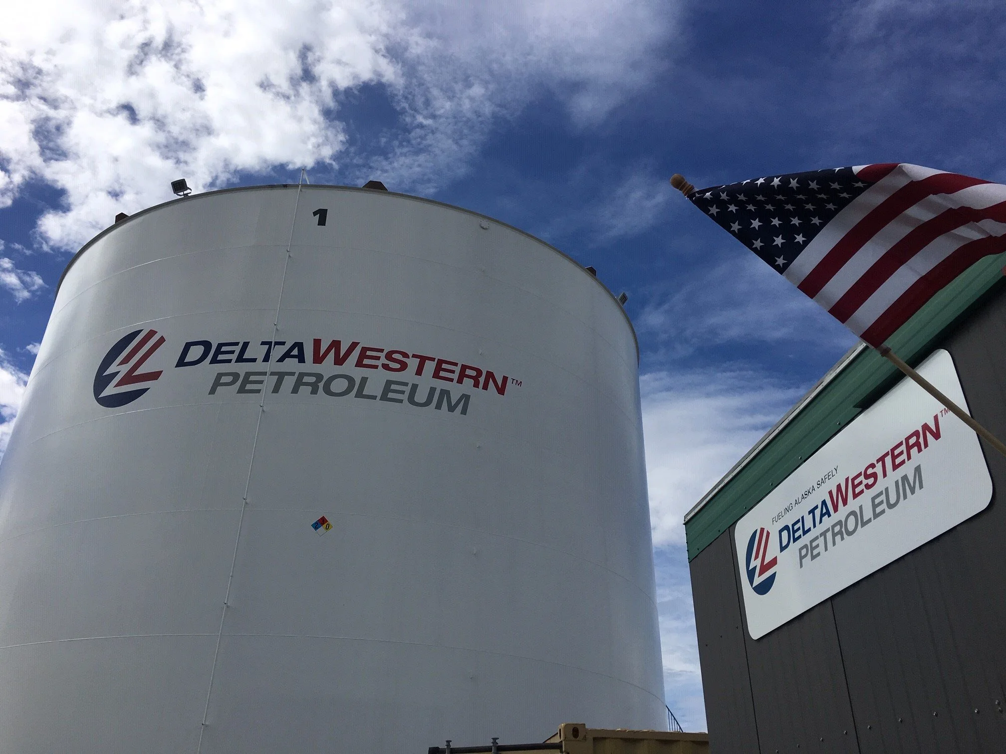 An industrial fuel storage tank with a Delta Western Petroleum logo, an American flag on a pole, and a sign indicating fuel safety regulations, against a partly cloudy sky.