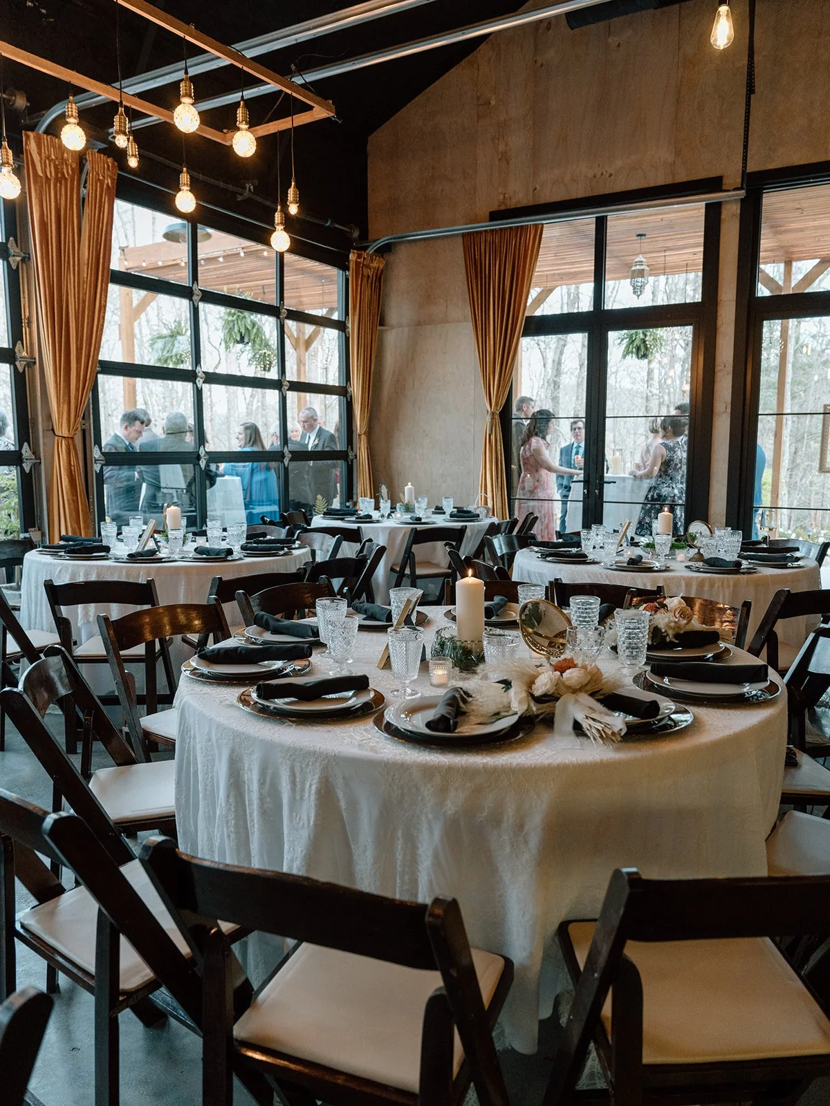 Elegant indoor dining area set for a celebration with multiple round tables, white tablecloths, black plates, napkins, and glassware, decorated with candles and floral arrangements, overlooking an outdoor patio with guests.