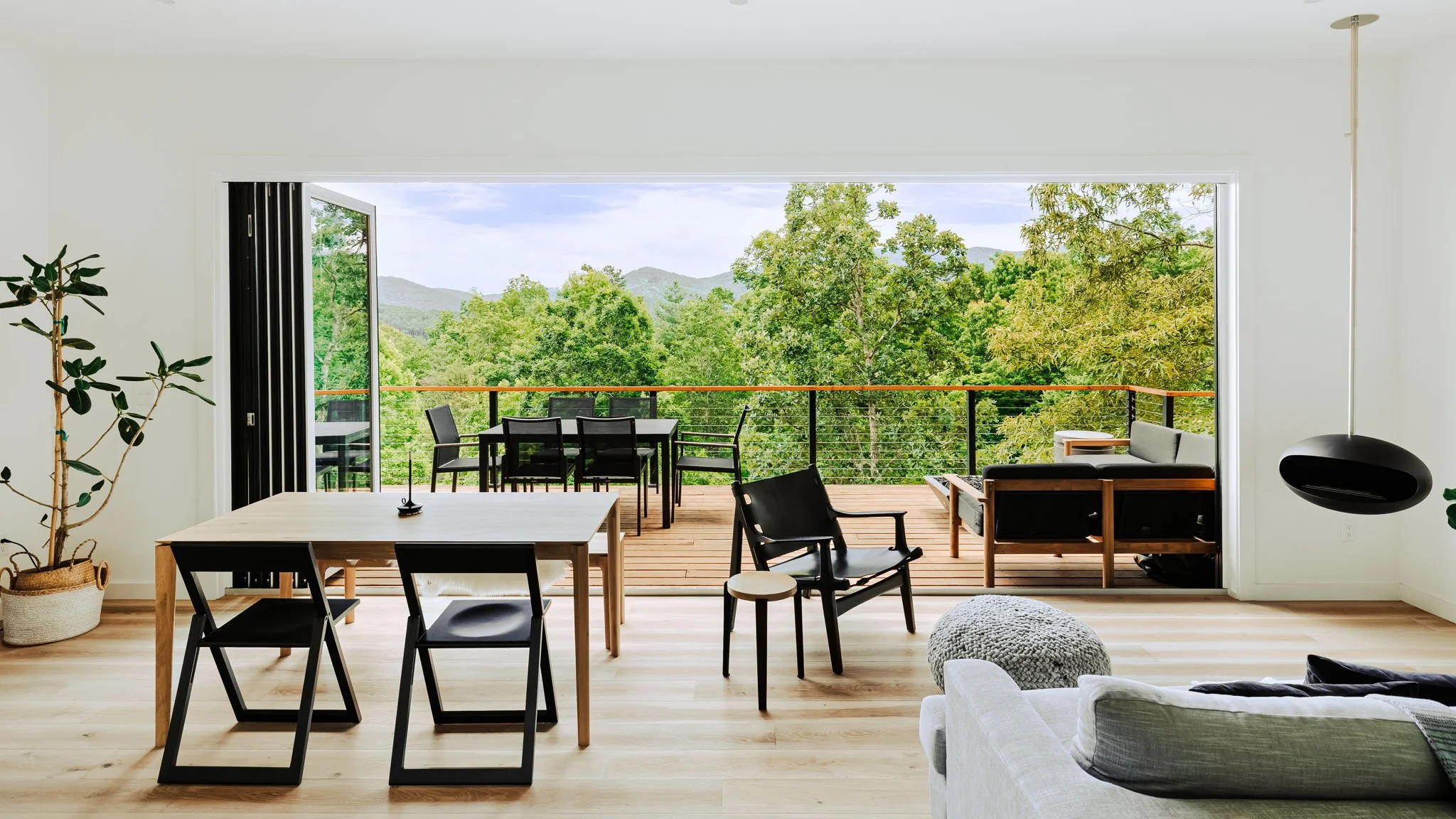 Living room with large open sliding door leading to a wooden balcony surrounded by green trees and mountains in the distance. Inside features a dining table with black chairs, some additional black chairs, a gray sofa with wooden armrests, and a potted plant.