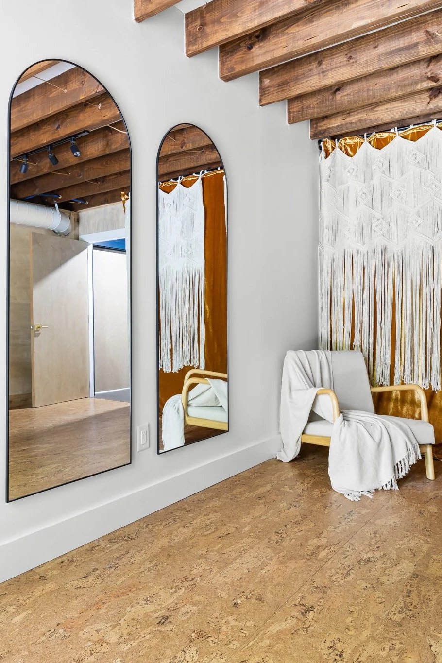 Interior of a modern room with two tall, arched mirrors on a white wall, a wooden sloped ceiling, a white armchair with a white blanket, and a decorative wall hanging with white and gold beads.