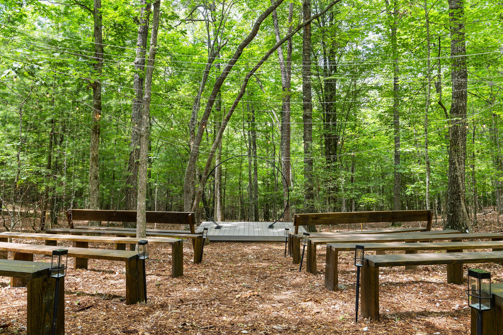 Wooden benches and a small deck with string lights, set in a lush green forest with trees and sunlight filtering through the leaves.