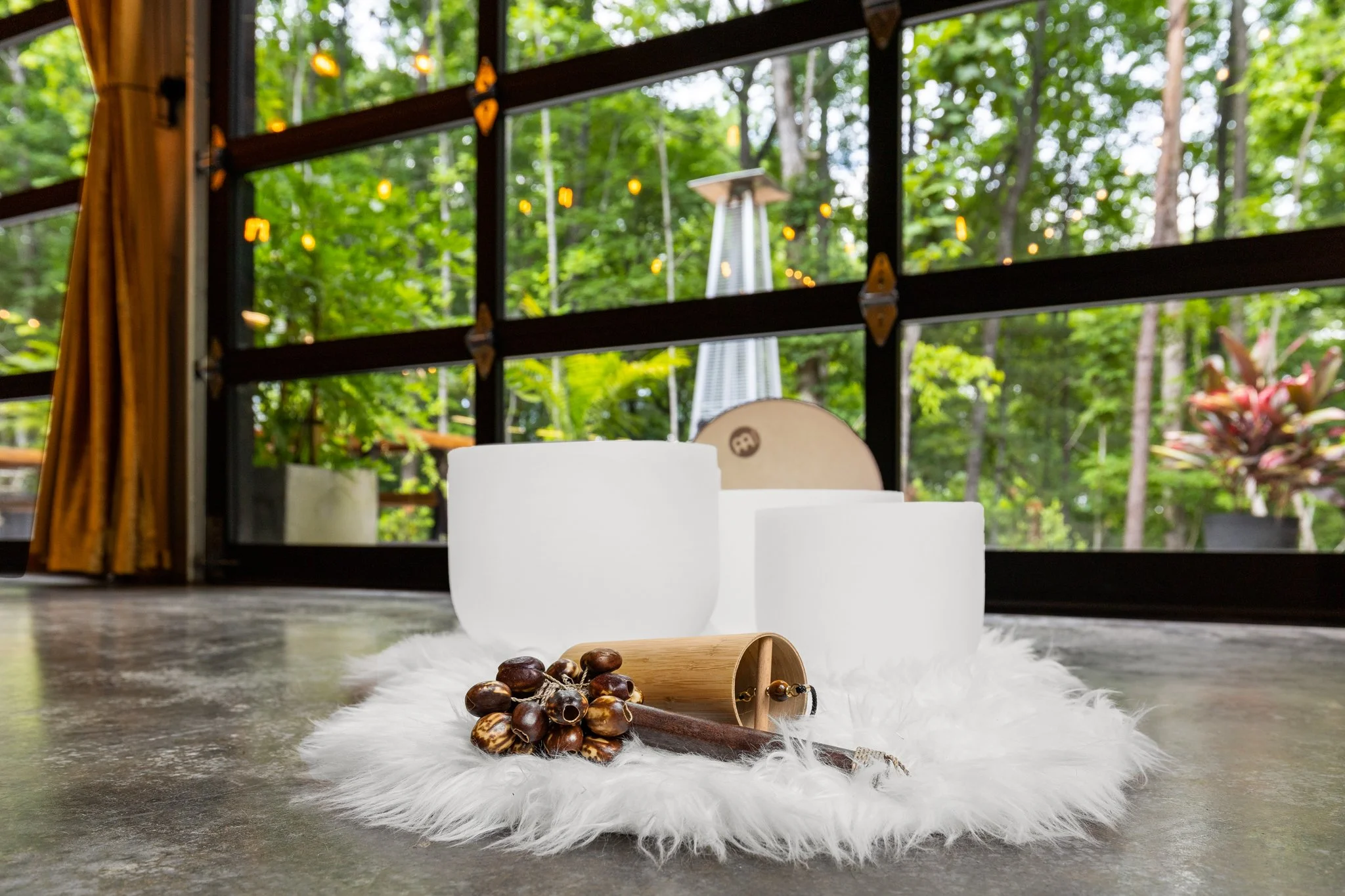 Indoor decor with white bowls on a fluffy white rug, small chestnuts, and a wooden scoop in the foreground. Large windows reveal green trees and outdoor string lights. Yoga retreat equipment. Sound bath singing bowls.