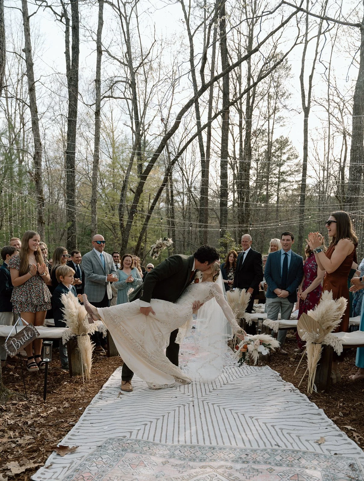 A bride and groom dancing or sparking a moment during an outdoor wedding ceremony in a forest setting, with guests watching and celebrating around them. The bride is wearing a white lace wedding dress, and the groom is in a suit, dipping her backward.
