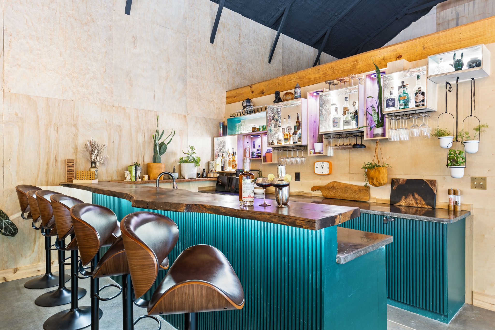 Modern bar or restaurant interior with a polished wooden bar counter, green and blue accents, and a wall decorated with potted plants, glassware, and liquor bottles.