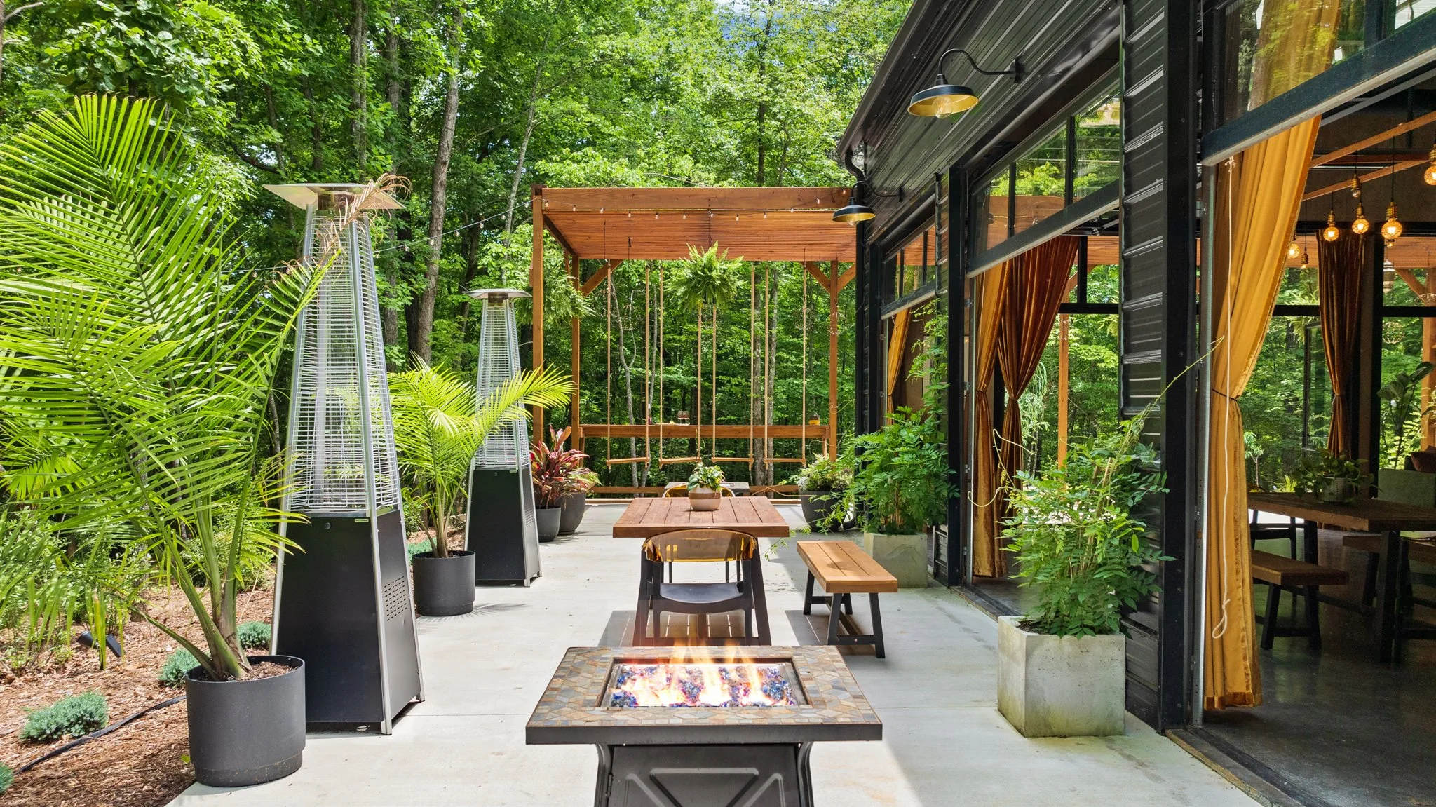 Outdoor patio with wooden furniture, potted plants, a fire pit, and a screened-in house with yellow curtains, surrounded by green trees.