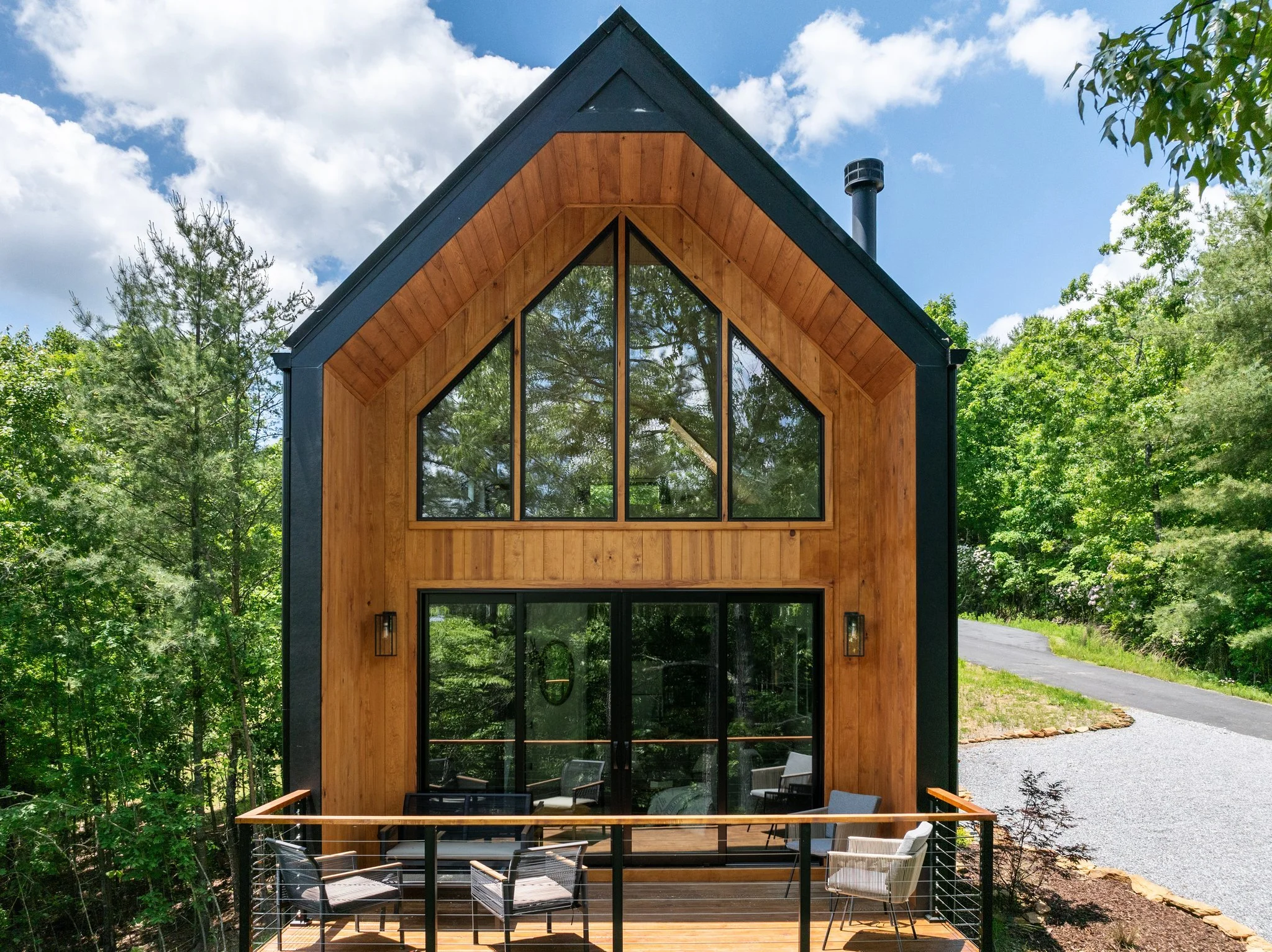 Modern house with large triangular windows, wooden exterior, surrounded by green trees, with a deck and outdoor seating area.