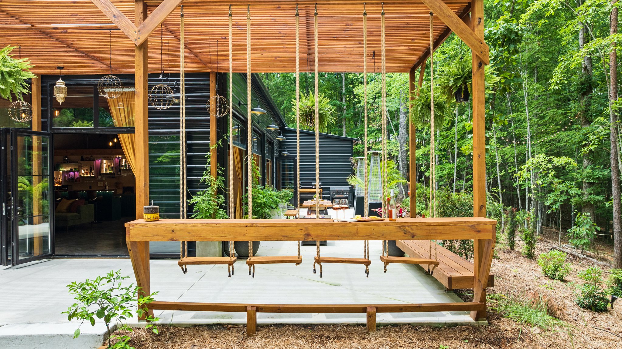 Outdoor wooden deck with a swing and string lights, surrounded by lush green trees and a black building with large glass doors and windows.