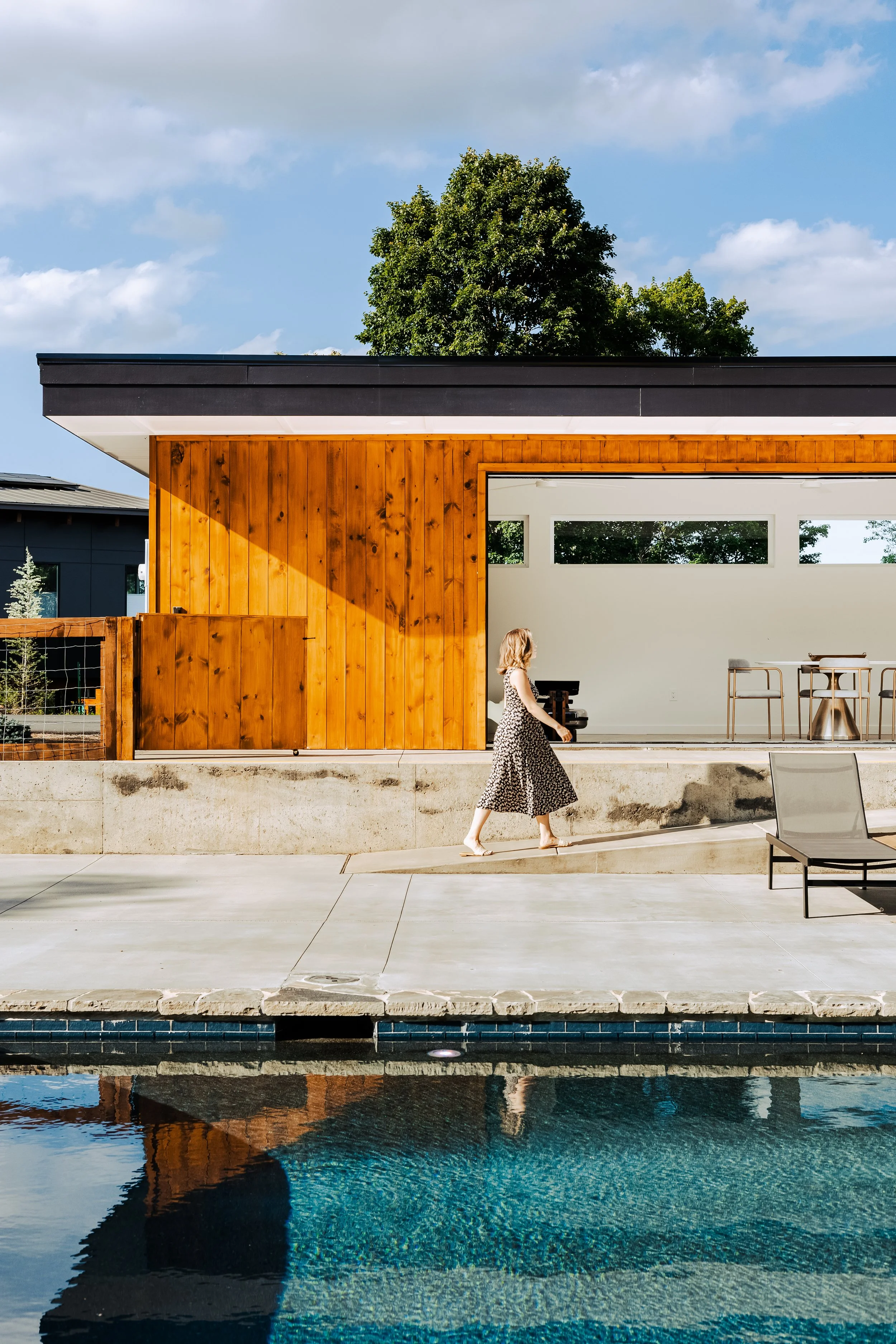 A woman in a long black and white patterned dress walks beside a modern house with a wooden exterior, large windows, and a black roof. There is a swimming pool in the foreground reflecting the house and sky, and the scene is under a partly cloudy sky.