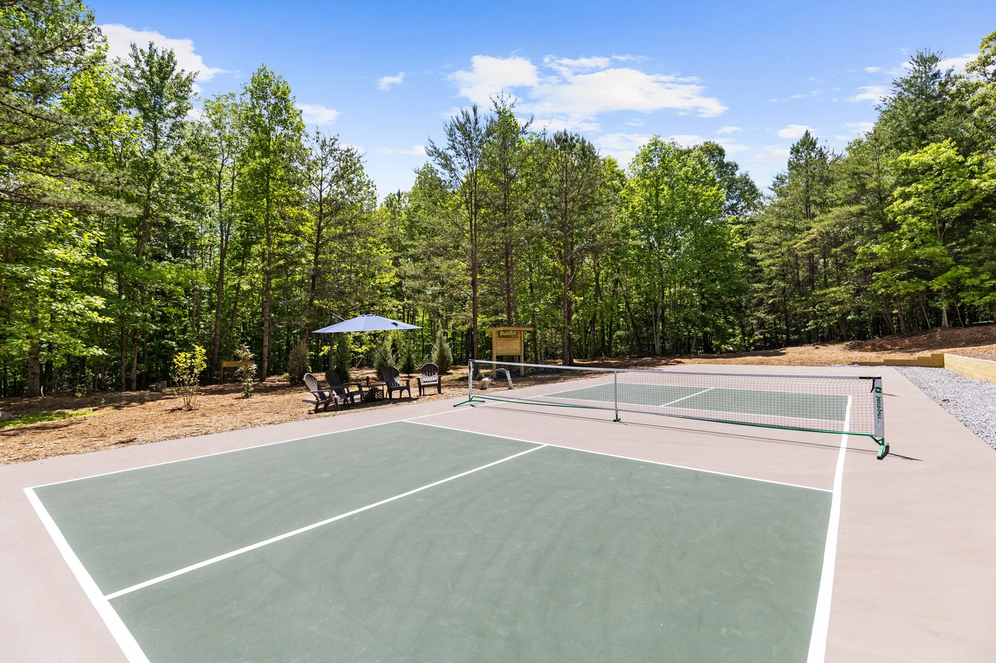 An outdoor pickleball court surrounded by trees with a seating area that includes chairs, an umbrella, and a volleyball net visible in the background.