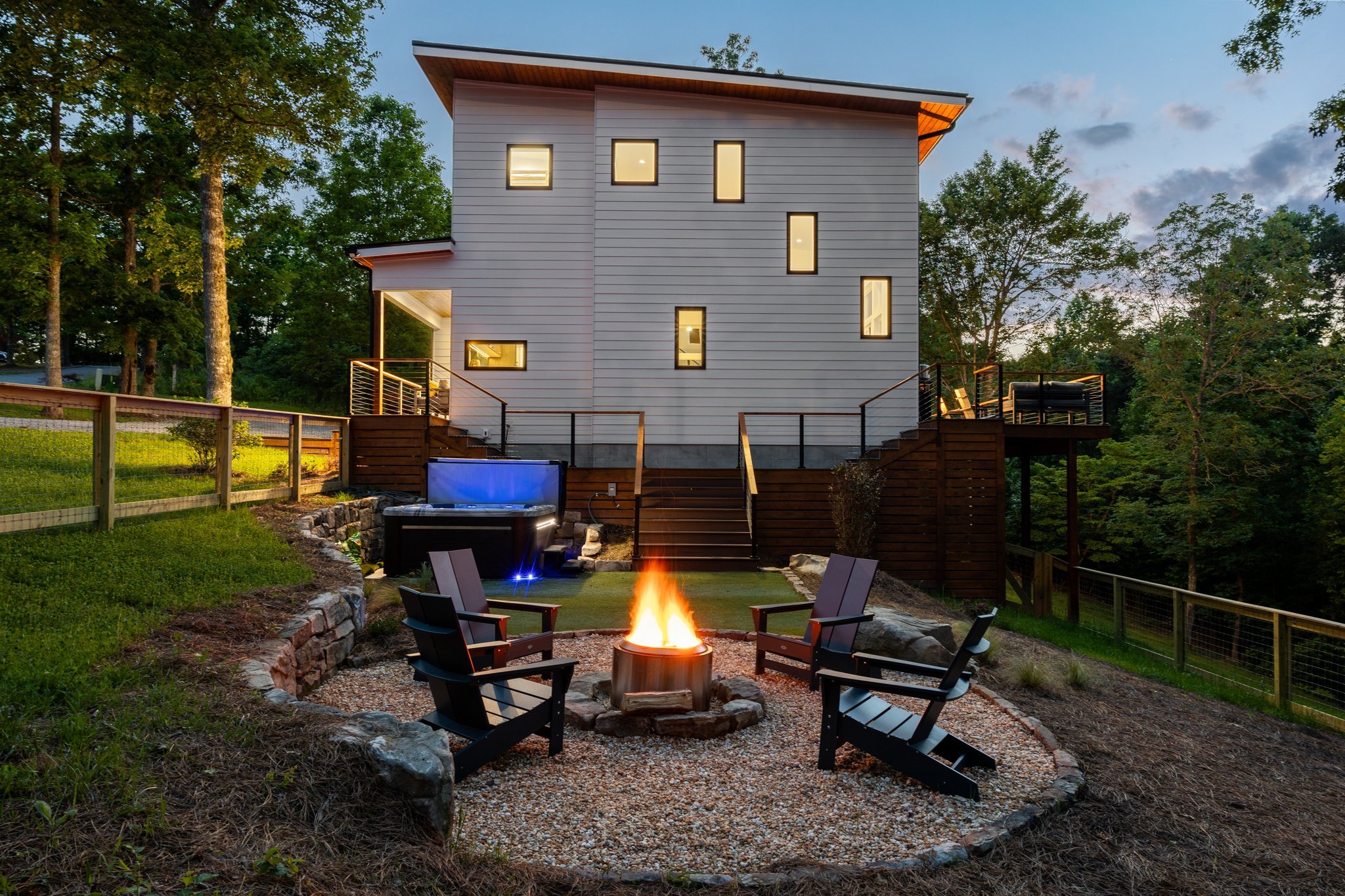 A modern house with multiple windows is illuminated at dusk and surrounded by trees. In the backyard, there is a fire pit with chairs around it and a small pool nearby. The porch area has outdoor furniture, and the scene is set for evening relaxation.