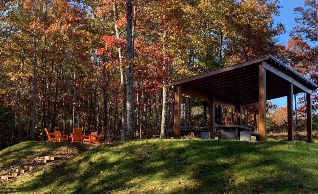 A grassy hill with a wooden pavilion and benches, surrounded by trees with colorful autumn leaves.