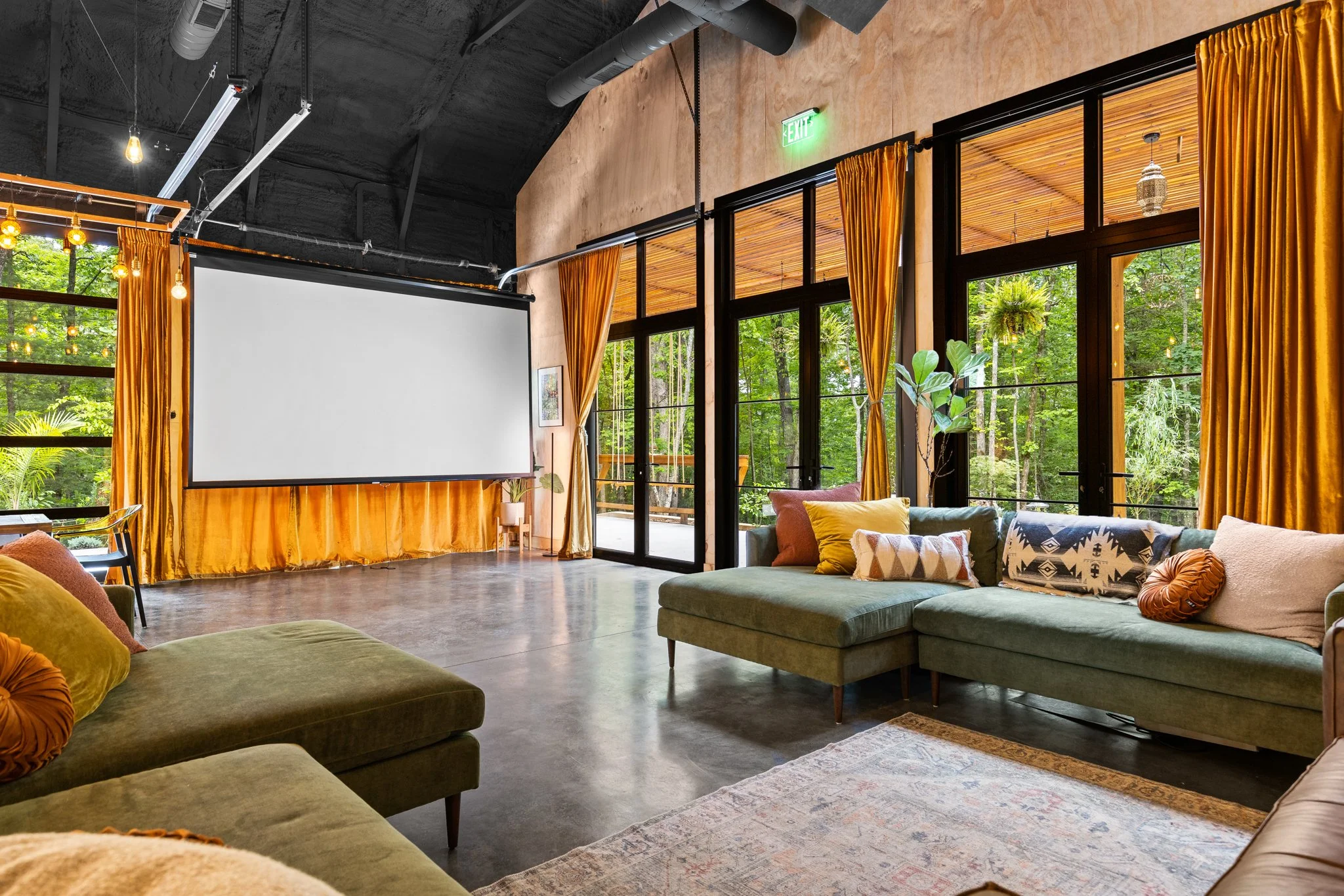 A cozy, modern living room with large windows, gold curtains, green and beige sofas with pillows, a rug, potted plants, and a projection screen.