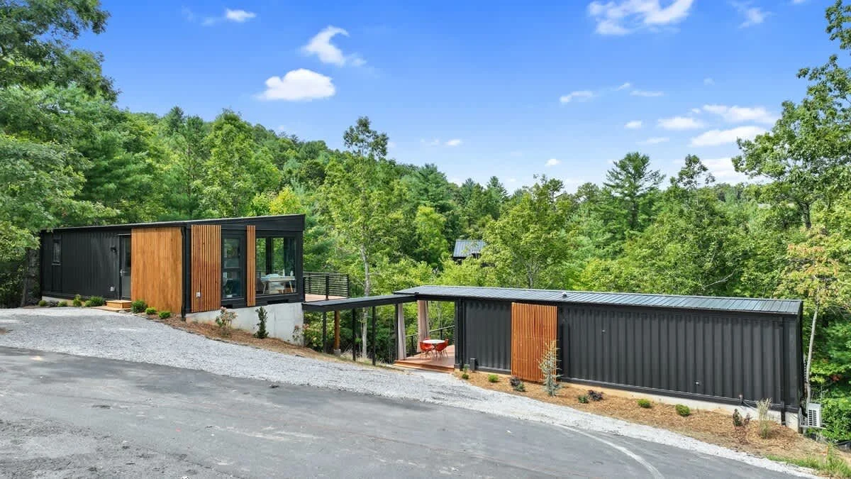 Modern black container homes with wood accents on a hillside surrounded by green trees under a blue sky.