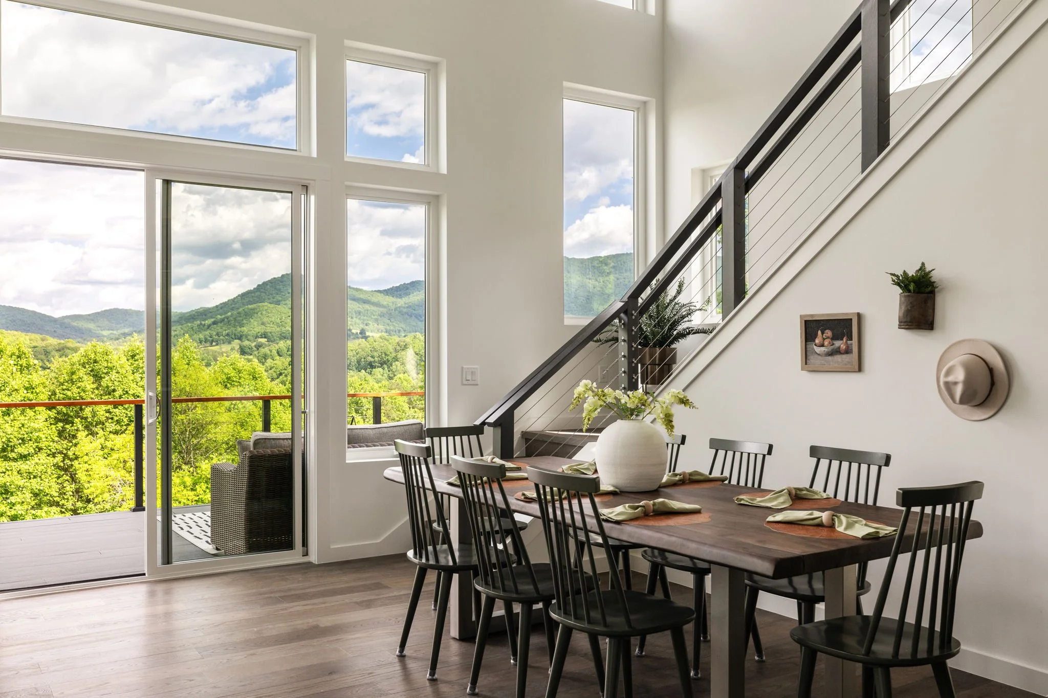 Modern dining room with large windows showing green hills and a partly cloudy sky, wooden dining table with chairs, a large white vase, potted plants, and wall art.