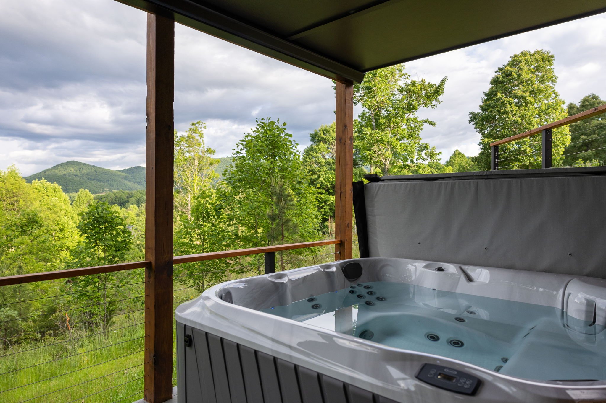 A hot tub filled with water on a balcony or deck with a view of green trees and mountains under a cloudy sky.