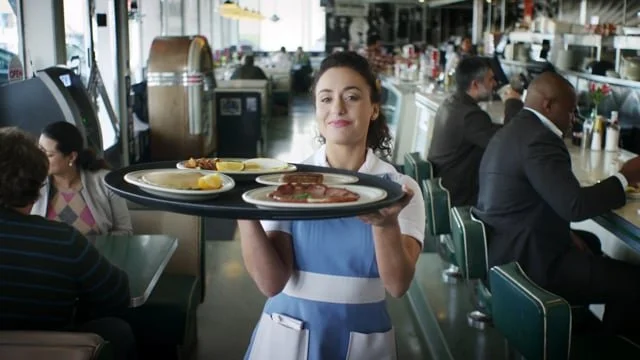 A waitress in a diner carrying a tray of breakfast items, including eggs, bacon, and pancakes.