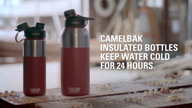 Two Camelbak insulated water bottles, one small and one large, on a rustic table in a cozy indoor setting. White text overlay reads: 'Camelbak insulated bottles keep water cold for 24 hours.'