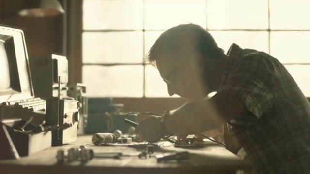 A man working at a cluttered desk in front of a window, focused on a task with various tools and electronic equipment around him.