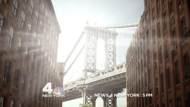 View of the Brooklyn Bridge in New York City, seen between brick buildings with a news banner at the bottom showing "NEWS 4 NEW YORK 5 PM."