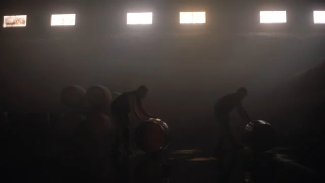 Silhouettes of people rolling barrels inside a dimly lit, large industrial space with rectangular windows.