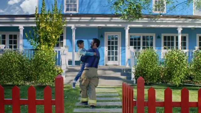 A firefighter holding a young boy in front of a large blue house with a porch, a red picket fence, and green bushes and trees.