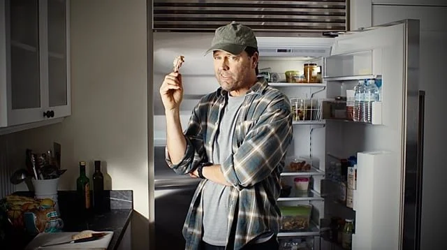 Man in casual clothing and cap eating a snack in front of an open refrigerator in a kitchen.