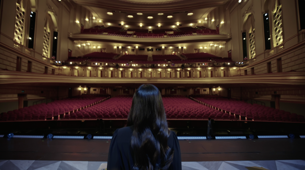 A woman with long dark hair stands on a stage in an empty, ornate theater, facing the audience seats and balconies.
