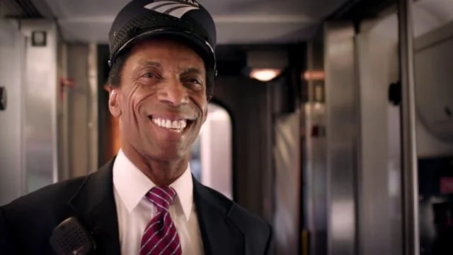 A smiling man in a conductor's uniform with a cap, standing inside a train.