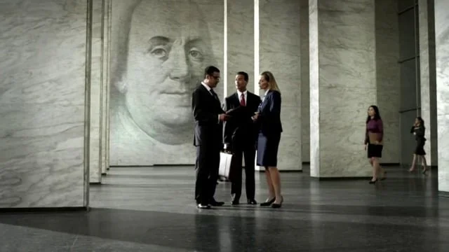Three business professionals in suits having a discussion in a spacious modern building with a large portrait of a man's face on the wall behind them, and two women standing apart in the background.