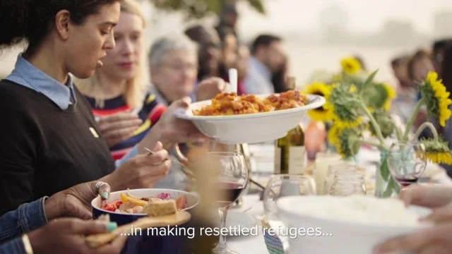People gathered at a long dinner table outdoors, eating and drinking, with flowers and sunlight.