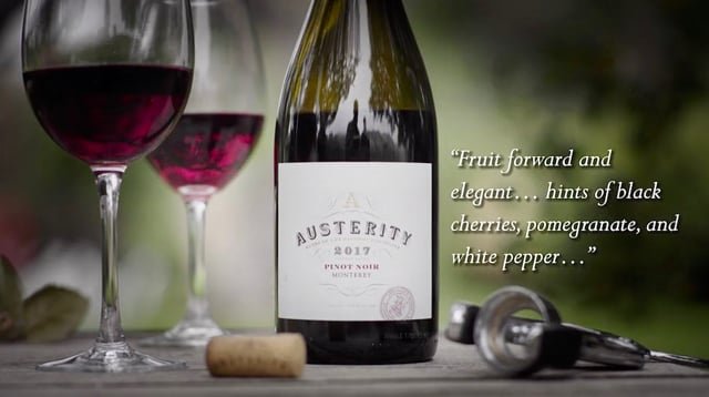 A bottle of wine labeled 'Austerity 2019 Pinot Noir,' two glasses of red wine, a wine cork, and a wine key on a wooden surface outdoors, with a quote about fruit, black cherries, pomegranate, and white pepper.