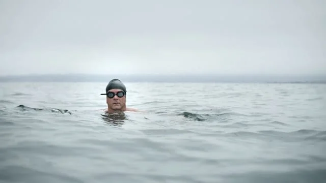 Swimmer in a gray swim cap and goggles in open water on a cloudy day.
