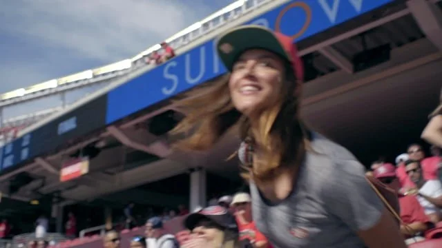 Smiling woman in gray T-shirt and red cap at a sports stadium with a crowd of spectators and a blue sign with white text in the background.