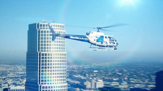 A helicopter flying near a tall skyscraper in a cityscape with a clear blue sky.